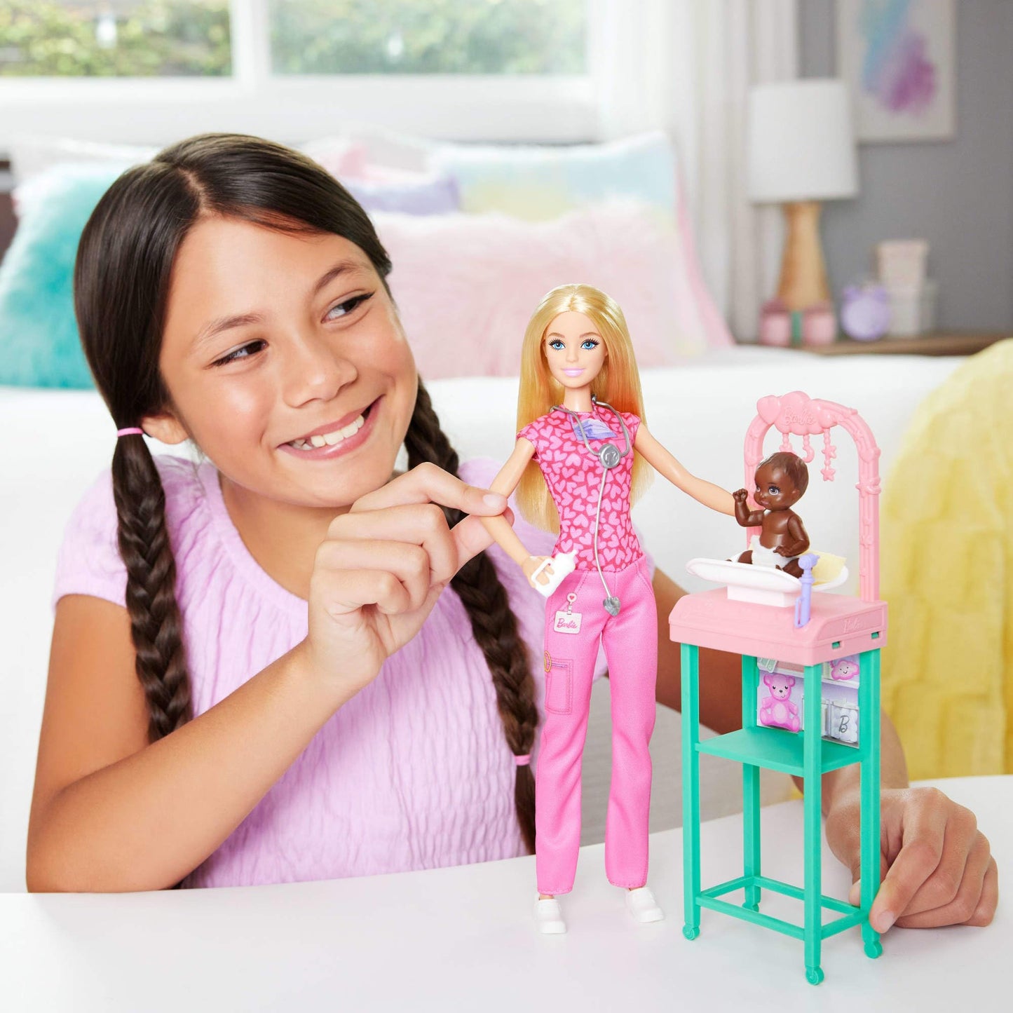 Barbie Baby Doctor doll with blonde hair wearing pink scrubs, includes baby accessories and furniture for imaginative play.