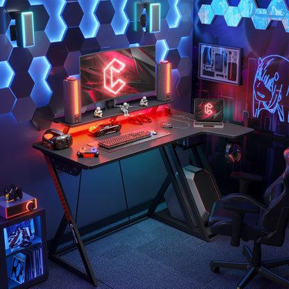Black CubiCubi L-shaped gaming desk with LED lights, monitor stand, shelves, hooks, and built-in power outlets for study and work.