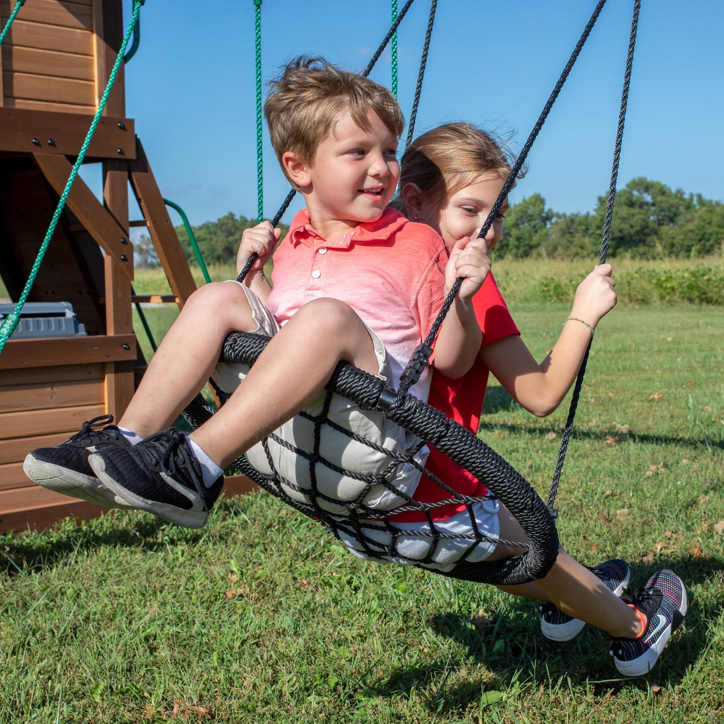 Backyard Discovery Cedar Cove cedar wood swing set with wave slide, tube slide, rock wall, web disc, and belt swings in a backyard.